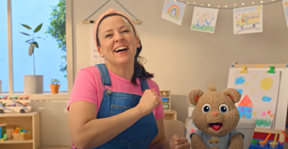 A woman laughs with a stuffed bear in a preschool classroom.