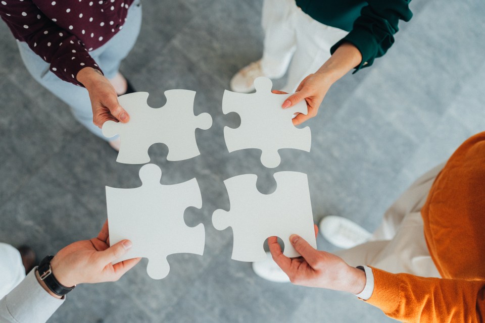Four people holding pieces of a white jigsaw puzzle.