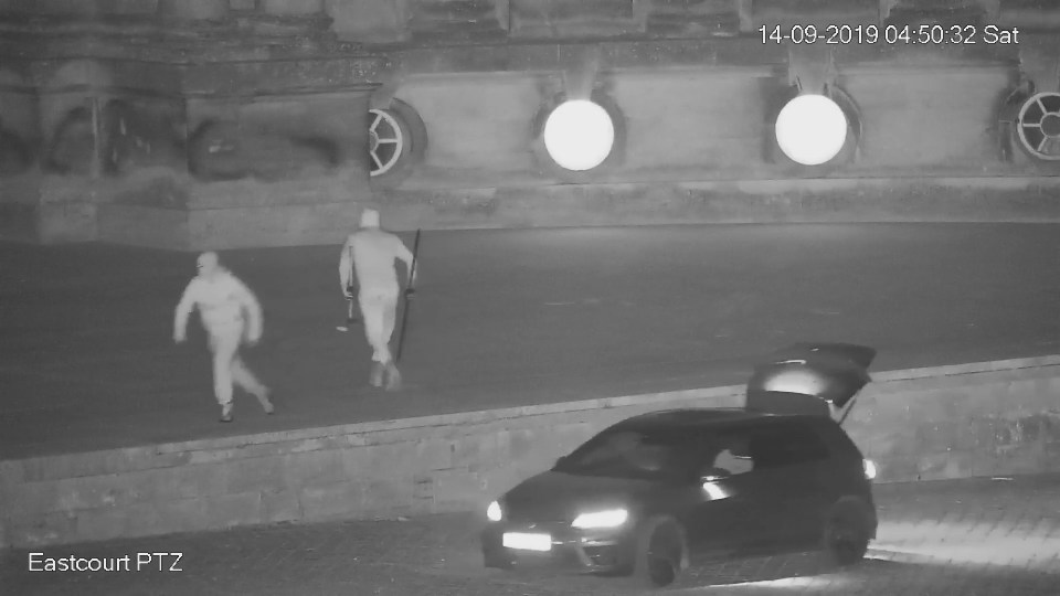 CCTV image of two men carrying a sledgehammer and crowbar near a car with its trunk open.
