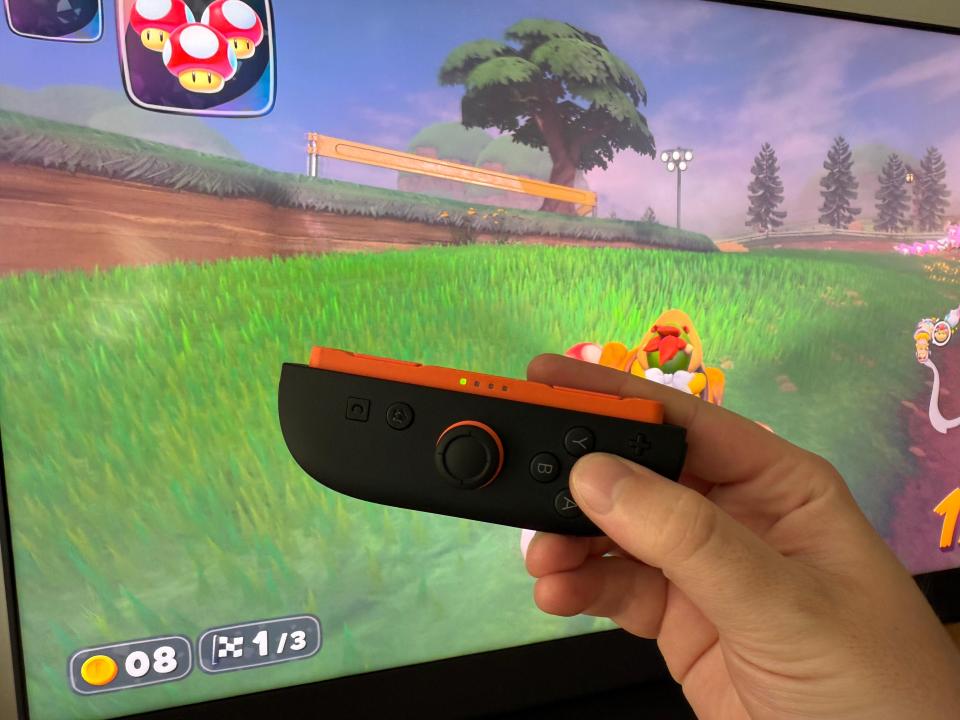 Hand holding a black and orange game controller in front of a screen showing a racing game.