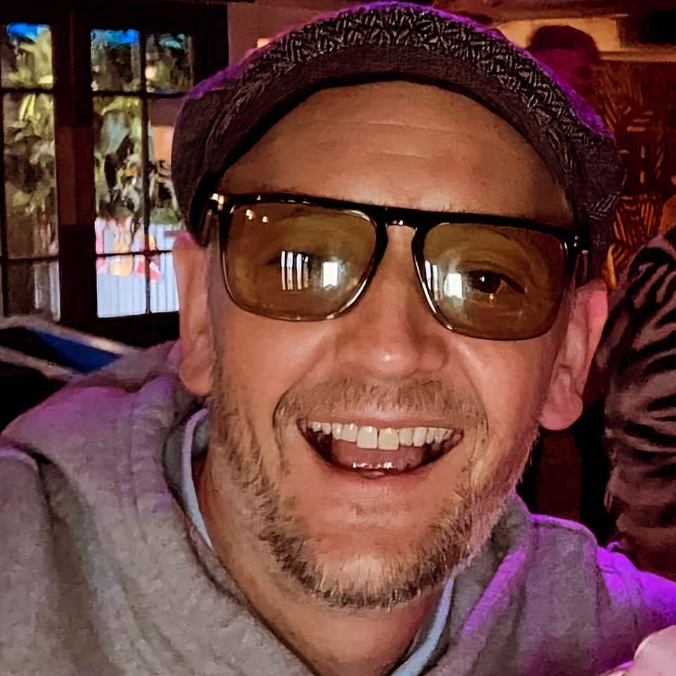 Close-up portrait of a smiling man wearing sunglasses and a cap.