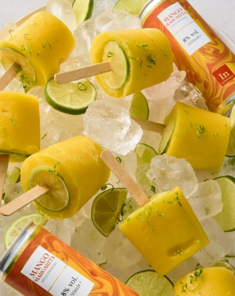 Mango margarita popsicles on a bed of ice with lime wedges and mango margarita cans.