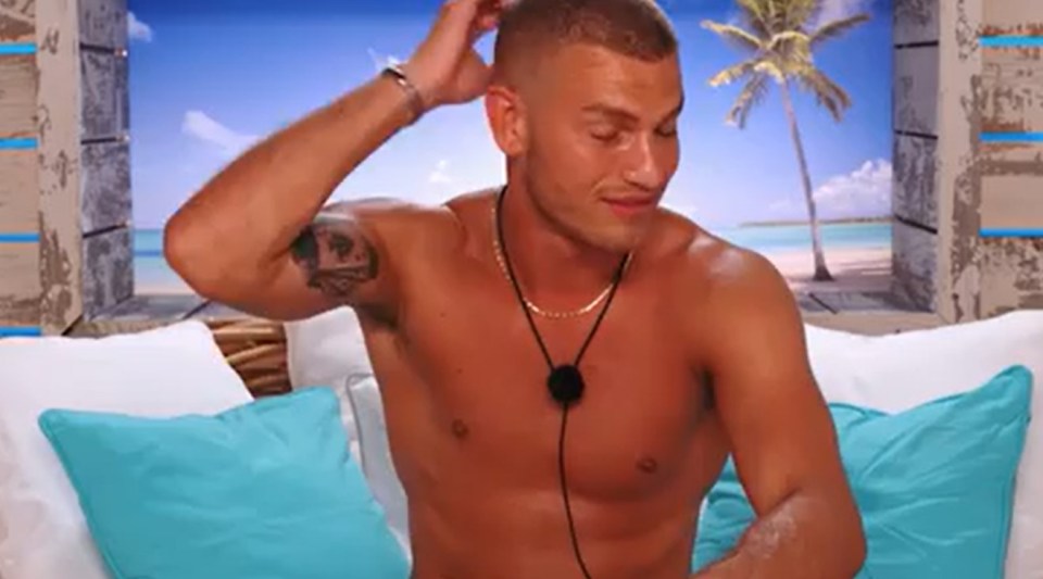 A shirtless man sits on a couch, scratching his head.
