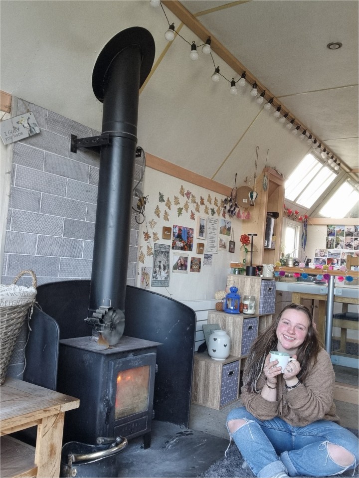 Woman sitting by a wood-burning stove in a cozy room.
