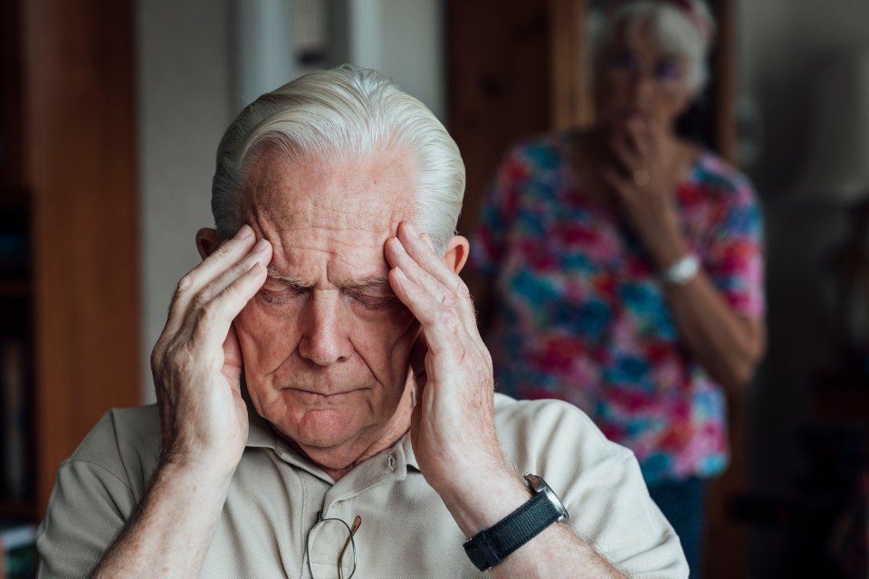 A senior man with Alzheimer's disease sits with his hands on his head, looking distressed; his wife is blurred in the background.
