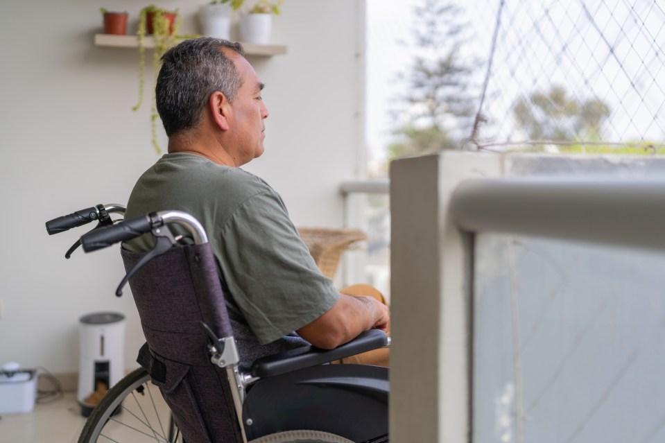 A man in a wheelchair sits pensively on his apartment balcony, looking out at the horizon.