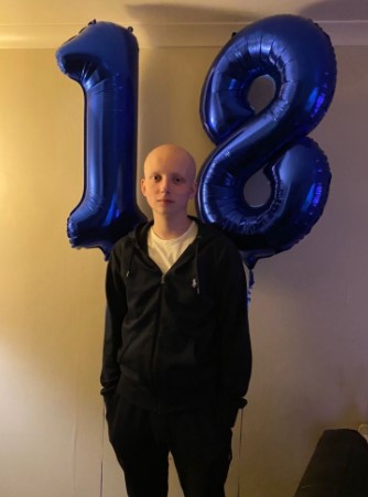 A young man with no hair stands in front of blue number 18 balloons.