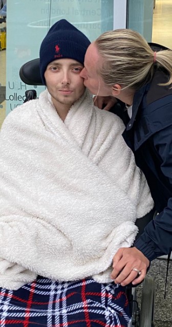 A terminally ill boy being kissed on the cheek by a woman, while wrapped in a blanket in a wheelchair.