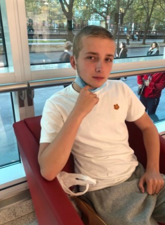 A teenage boy sits in a red chair, adjusting his face mask.