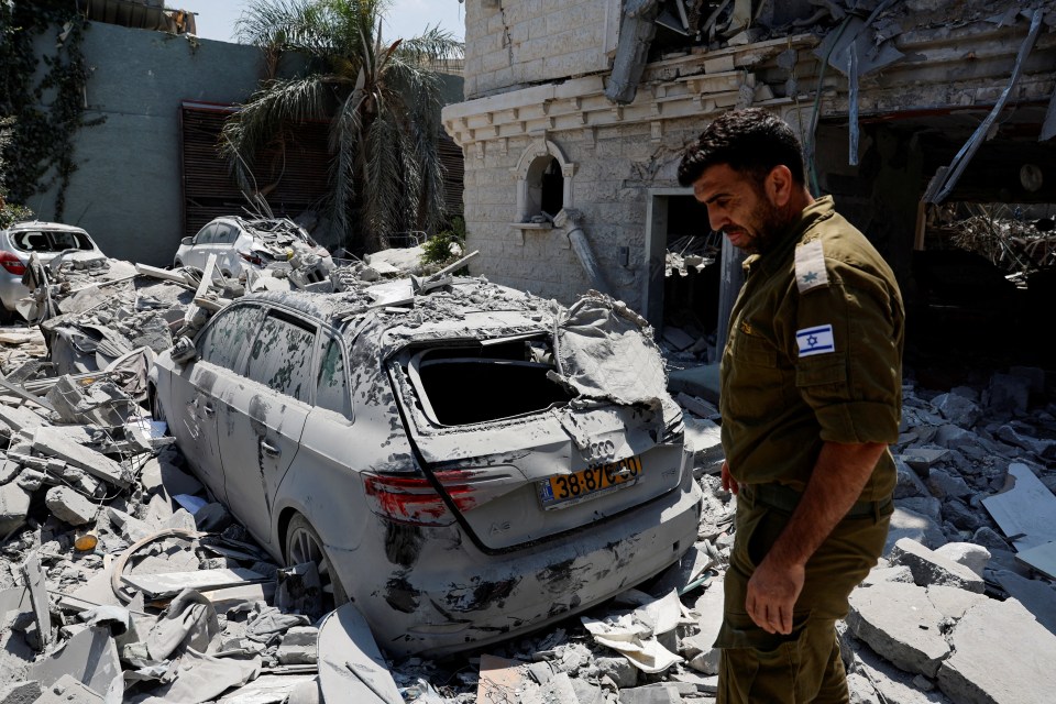 Israeli soldier near damaged vehicles and buildings after missile attack.