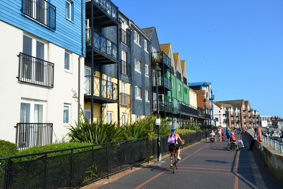 Modern seafront apartments in Littlehampton, England, with people walking and cycling along a paved path.