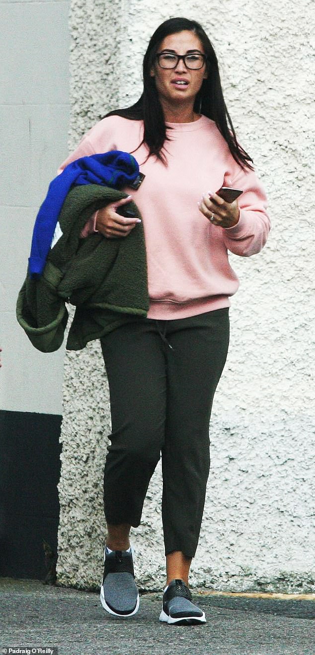 Charlene holds her phone as she walks in grey trainers, dark green joggers and a pink sweater