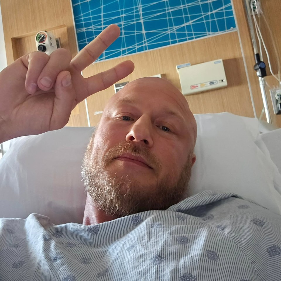 Bald man in a hospital bed giving a peace sign.