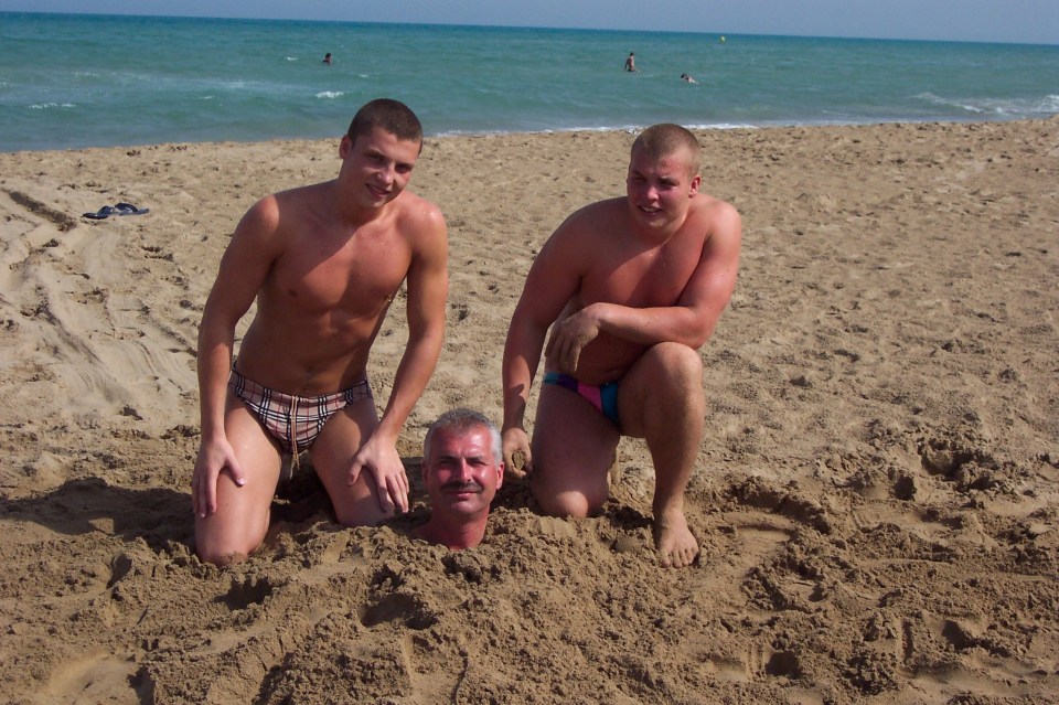 Man buried in sand on beach with two men kneeling beside him.
