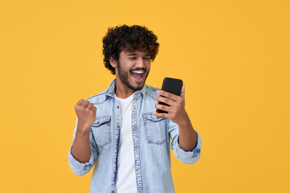 A joyful young man celebrates on his phone against a yellow background.