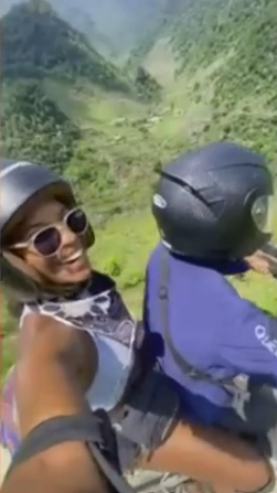 Screenshot of a woman smiling and taking a selfie while riding on the back of a motorcycle in a mountainous area.