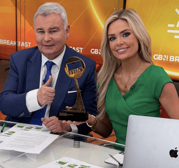 Two news anchors at a desk, one holding an award and giving a thumbs up.