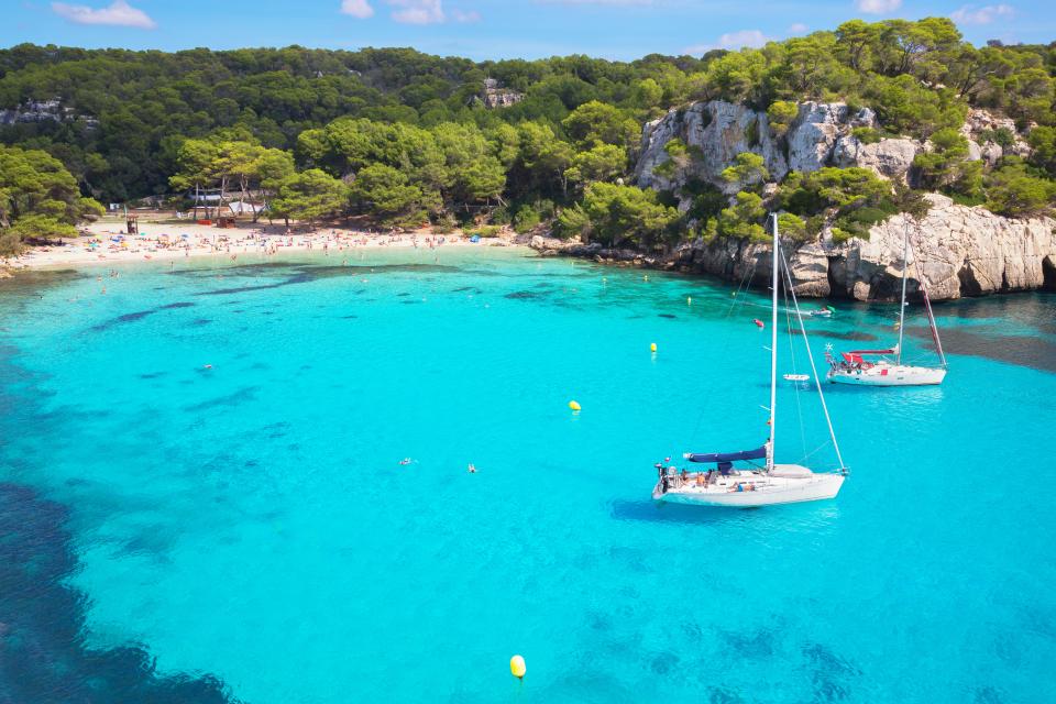 Aerial view of Cala Macarella beach in Menorca, Spain, with sailboats in turquoise water.