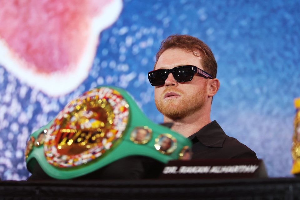 Canelo Álvarez at a press conference, wearing sunglasses and holding a championship belt.