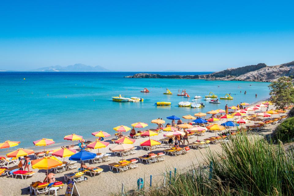 Paradise Beach in Kefalos, Kos, Greece with colorful umbrellas and beachgoers.