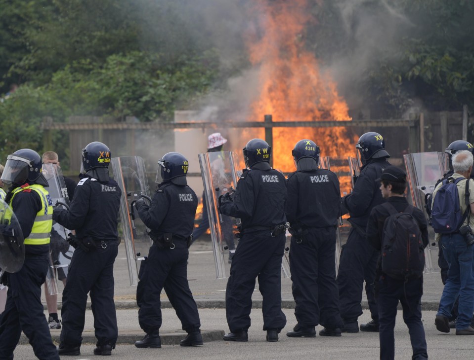 Riot police standing behind shields during a demonstration with a fire in the background.