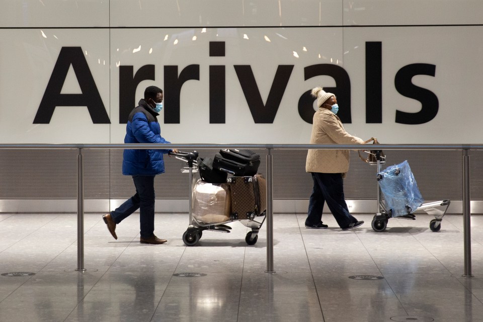 Two masked passengers with luggage carts walk past an arrivals sign at an airport.