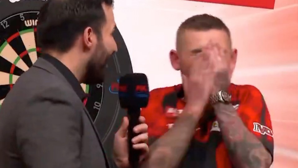 A post-game interview with a darts player covering his face with his hands while an interviewer holds a microphone.