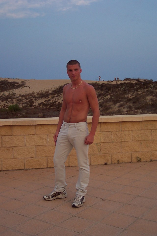 Shirtless man in light-colored pants standing on a patio with a sand dune in the background.