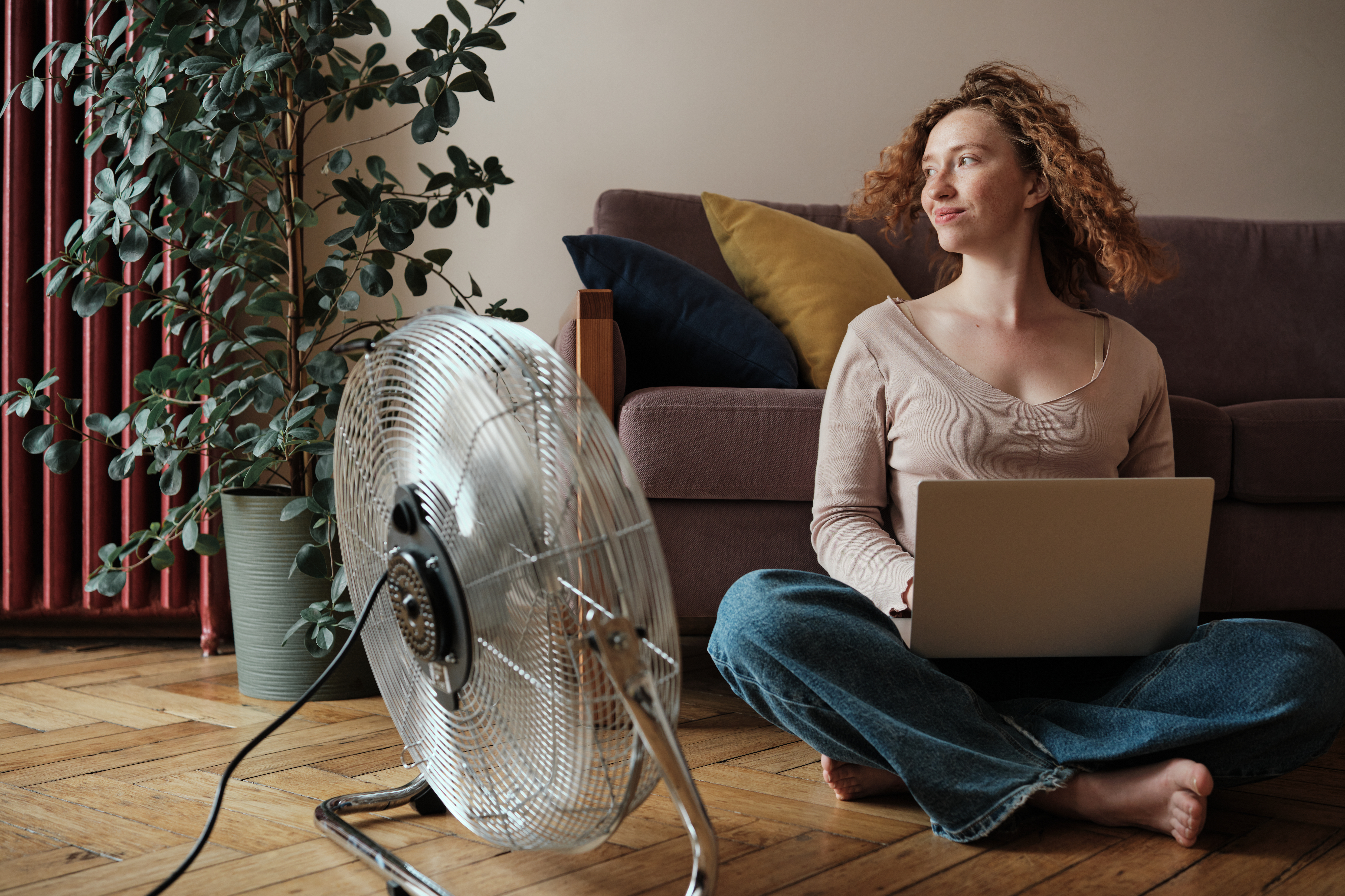 Woman freelancer using laptop while sitting on floor near electric fan, working from home during summer heat