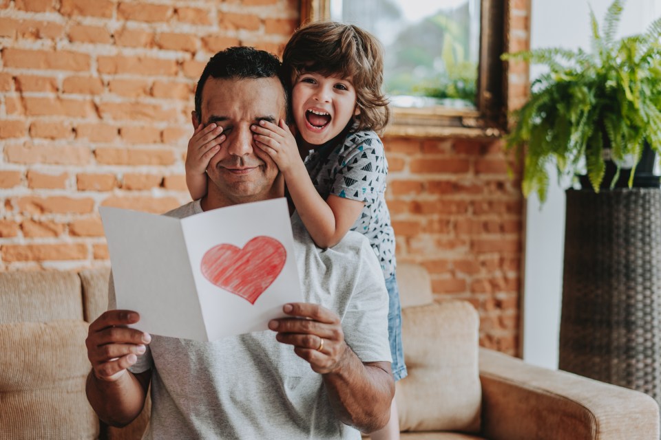 A child surprises his father with a handmade card featuring a heart.