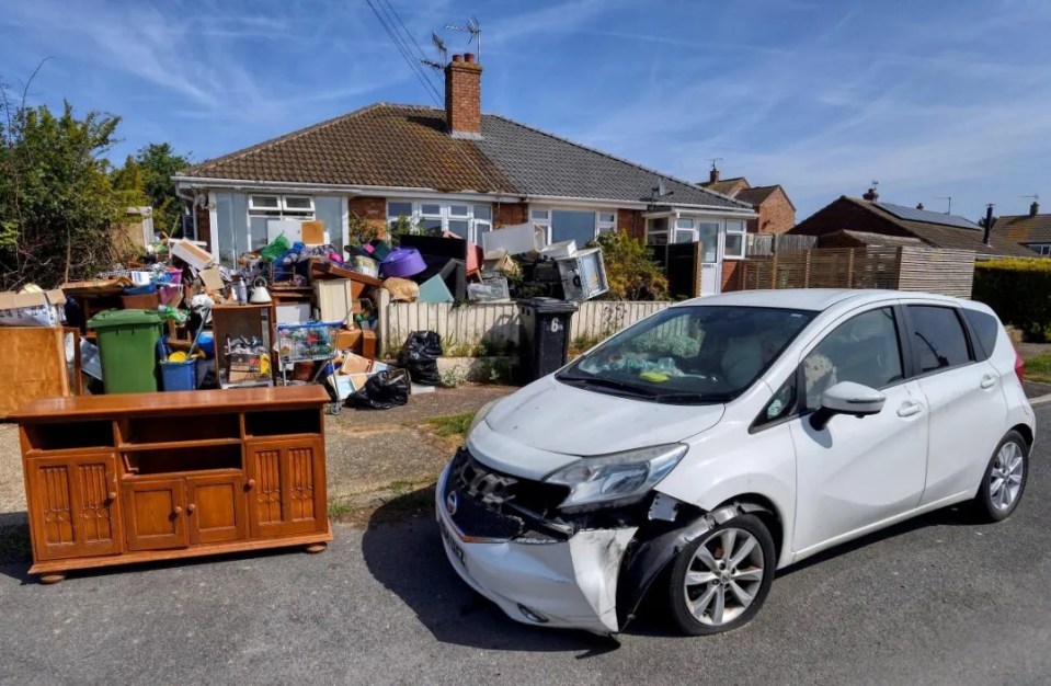 A pile of discarded furniture and household items sits in front of a house, next to a damaged white car.