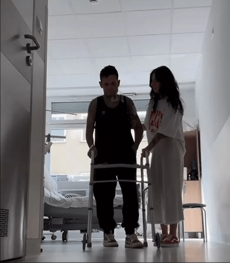 Man using a walker in a hospital hallway, assisted by a woman.