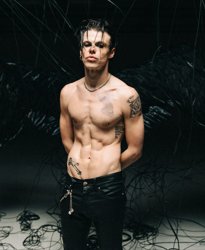 Yungblud, shirtless, with tattoos, standing amidst tangled wires.