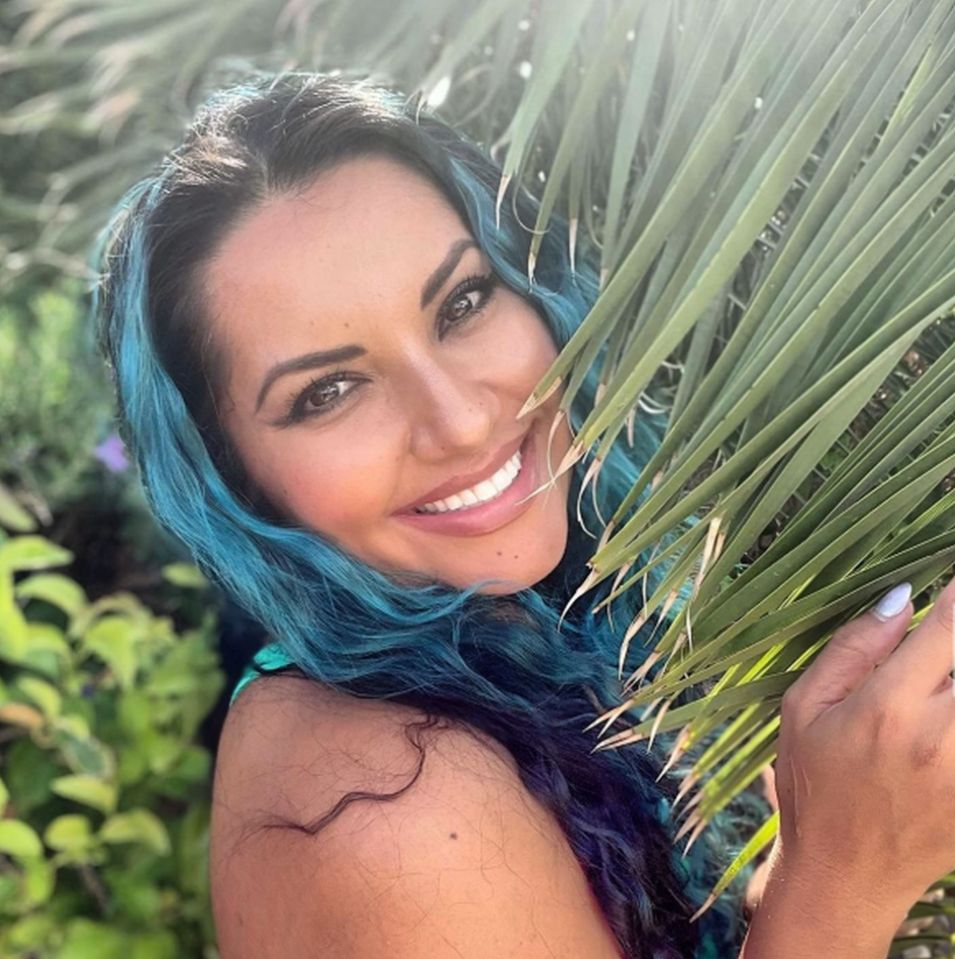 Woman with teal hair partially obscured by palm fronds.