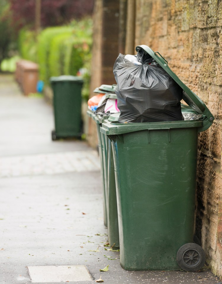 Several green wheelie bins overflowing with black garbage bags on a pavement.