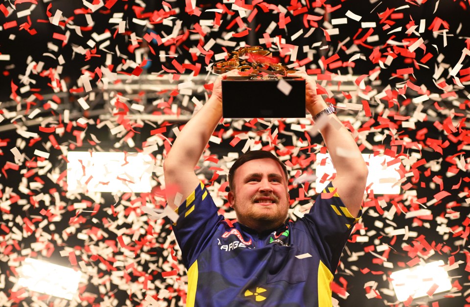 A darts player raises the Ladbrokes UK Open trophy amidst falling confetti.