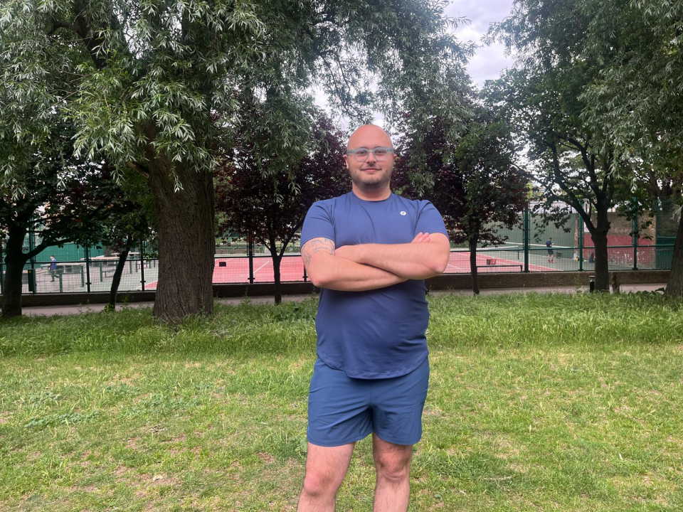 Man standing with arms crossed in a park near a tennis court.
