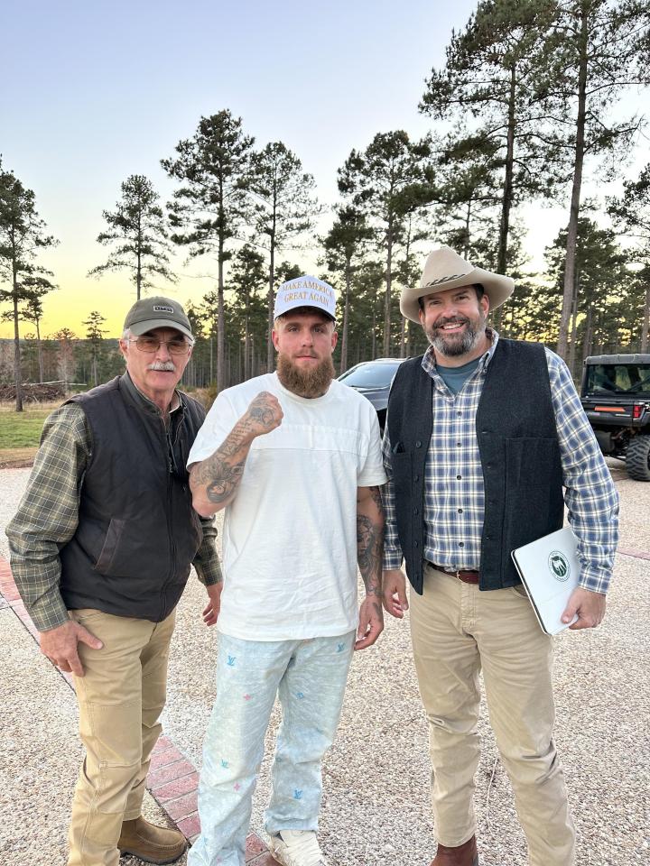 Three men standing outdoors in front of trees.  The central man wears a "Make America Great Again" hat.