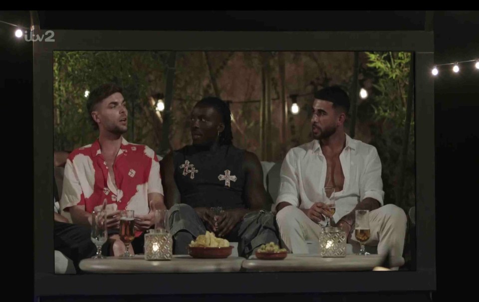 Three men relaxing outdoors, watching a large screen.