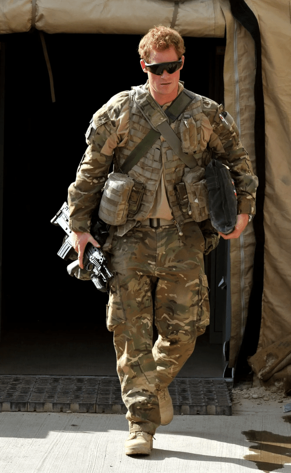 Prince Harry in military uniform carrying a rifle and bag.