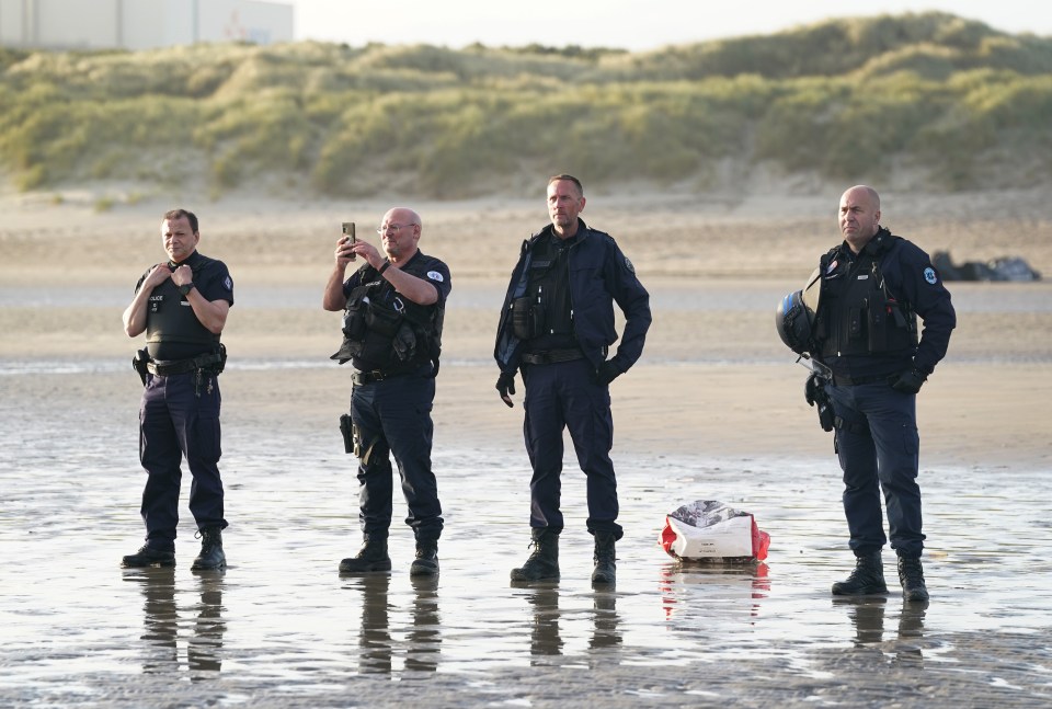 French police officers on a beach watch a group of people board a small boat.