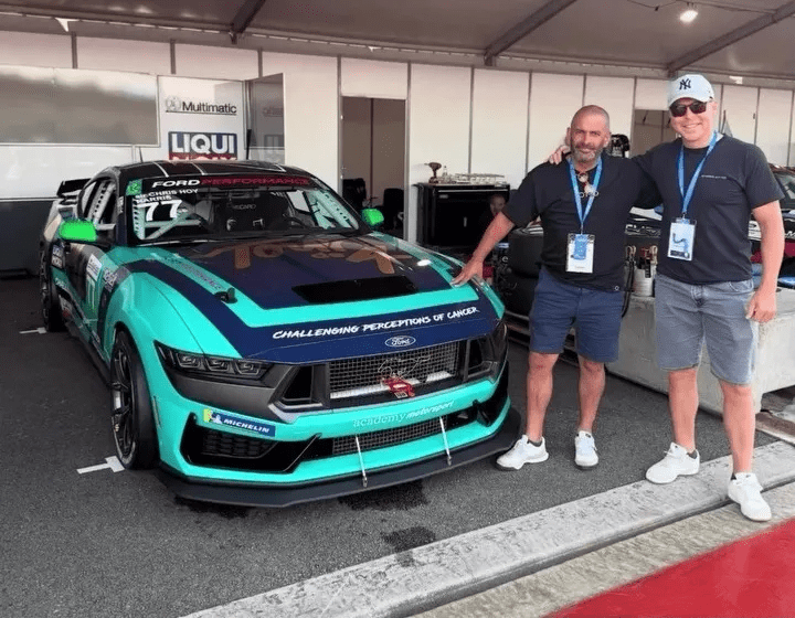Two men stand beside a teal Ford Mustang race car.