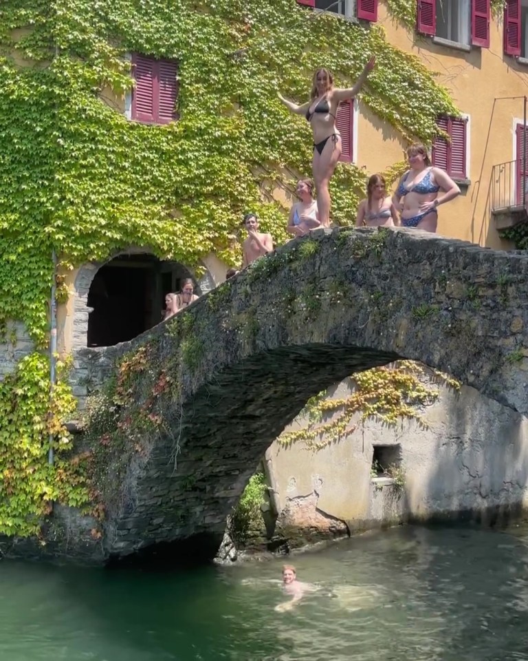 A group of women on a stone bridge over water, one woman is poised to jump. A person is swimming below.