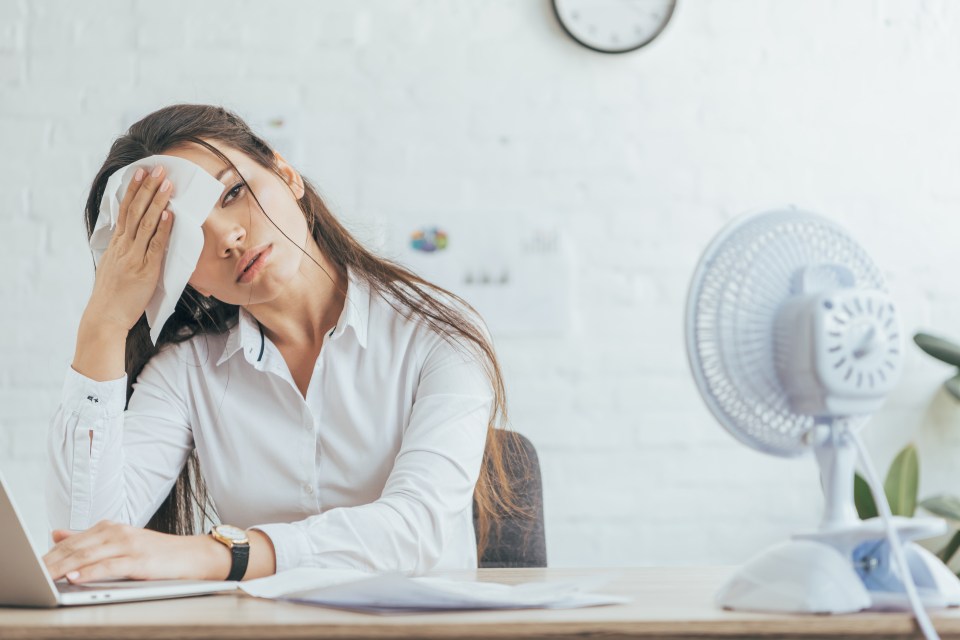 Sweaty businesswoman wiping her forehead while working at her desk with a fan nearby.