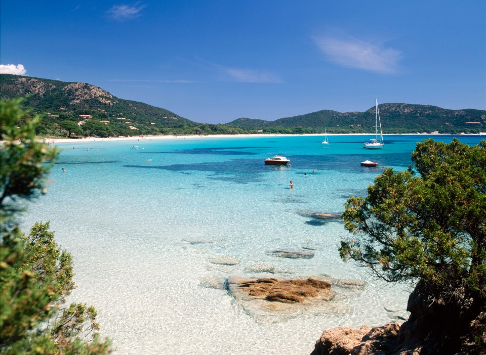 Palombaggia Beach in Corsica, with clear turquoise water, boats, and people swimming.