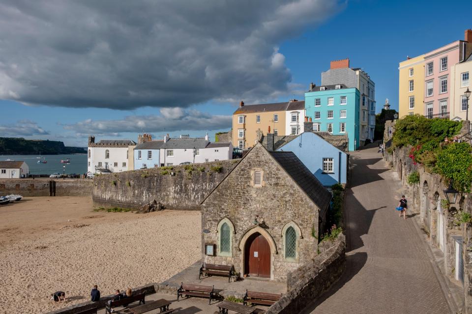 View of Penniless Cove Hill in Tenby, Wales, showing colorful houses, a stone building, and a sandy beach.