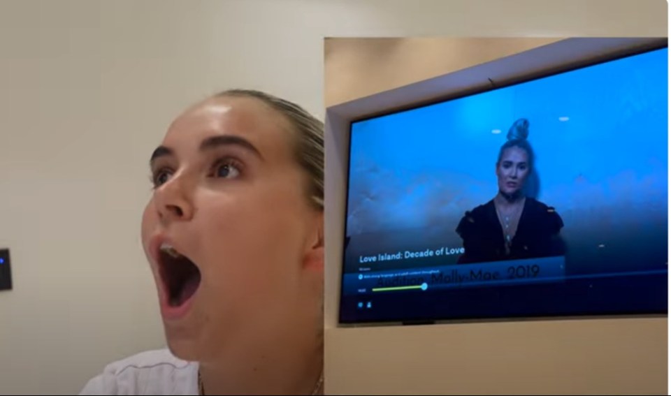 A woman reacting with a surprised expression next to a TV screen showing a video titled "Love Island: Decade of Love" by Molly-Mae.