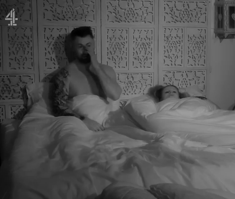 Man on phone in bed next to sleeping woman.