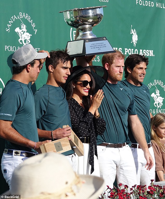 'As the trophy is raised, Meghan is pushed to the back and squashed under the cup, needing to hold her large-brimmed hat until she manages to move forward to perform some seal-claps of congratulation,' says Ms James