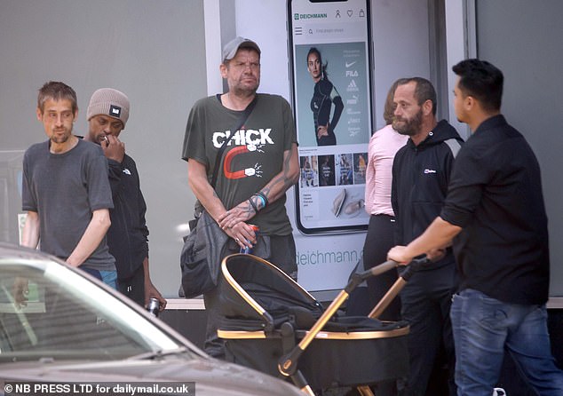 A man with a beer can loiters in a shop doorway as a local shopper walks past pushing a baby in a pram